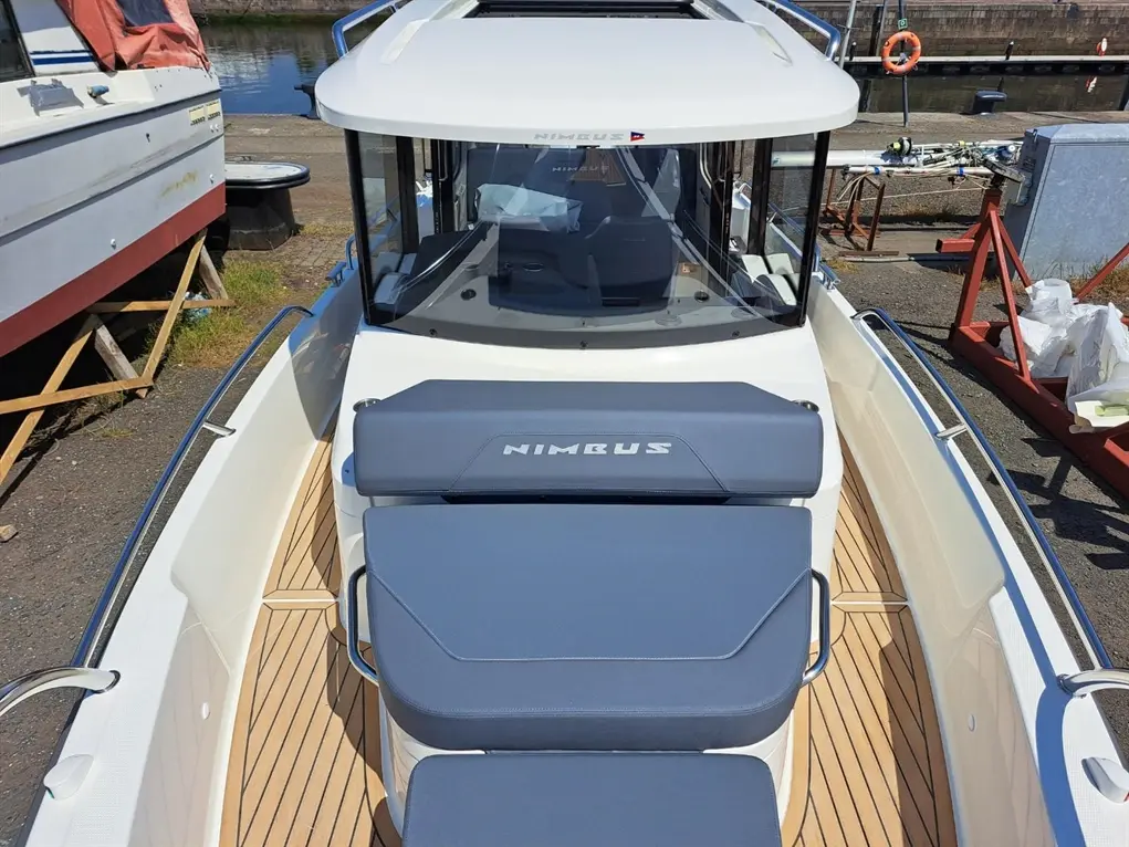 Nimbus boat interior with a sunbathing area, blue cushions, and a gleaming wooden deck by the water, showcasing nautical design.